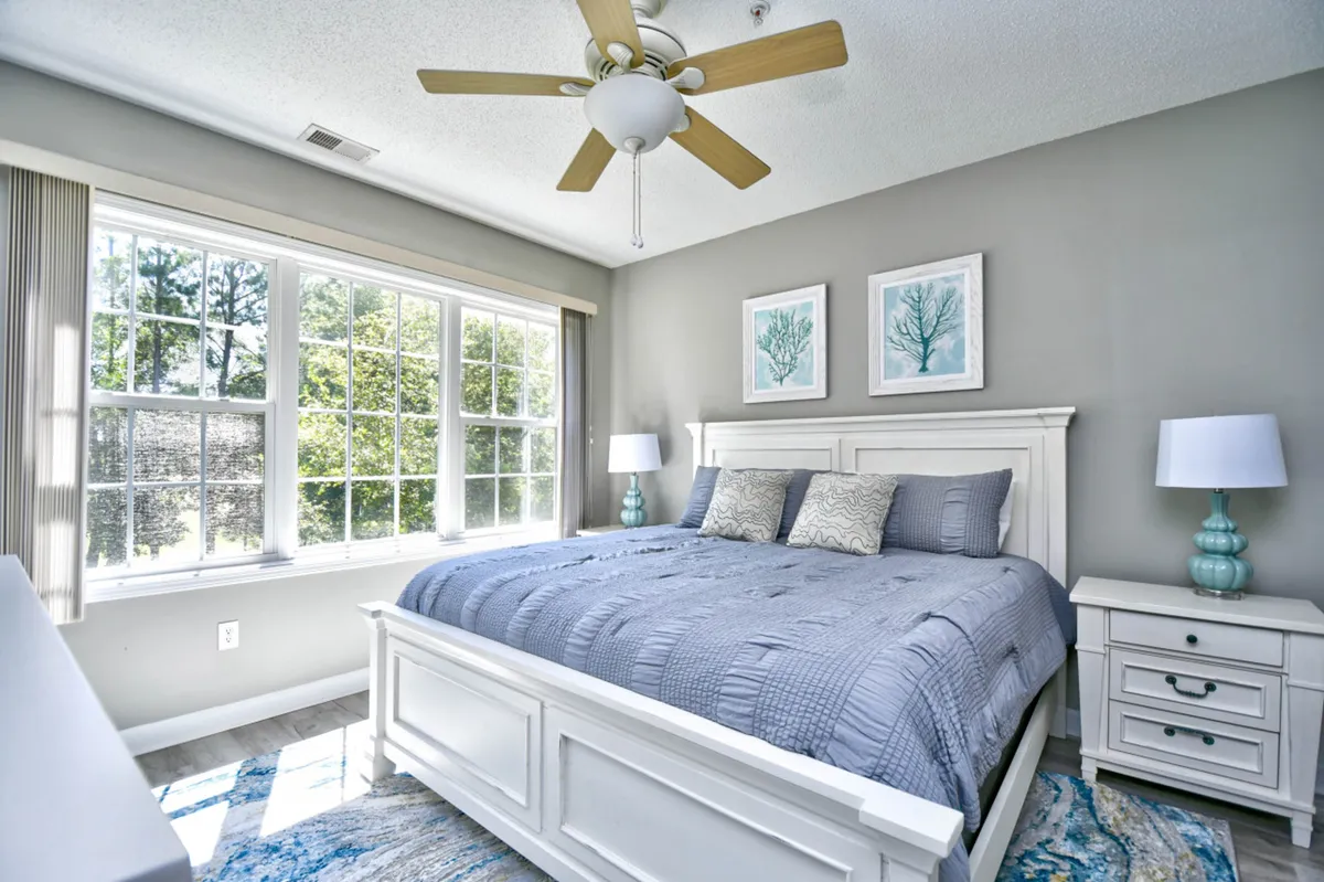 The image showcases a serene bedroom with a plush bed, large sunlit windows, tasteful coastal decor, and a cozy nightstand, evoking comfort and relaxation. Natural light fills the space, inviting guests to unwind, read by the window, or wake up rejuvenated.