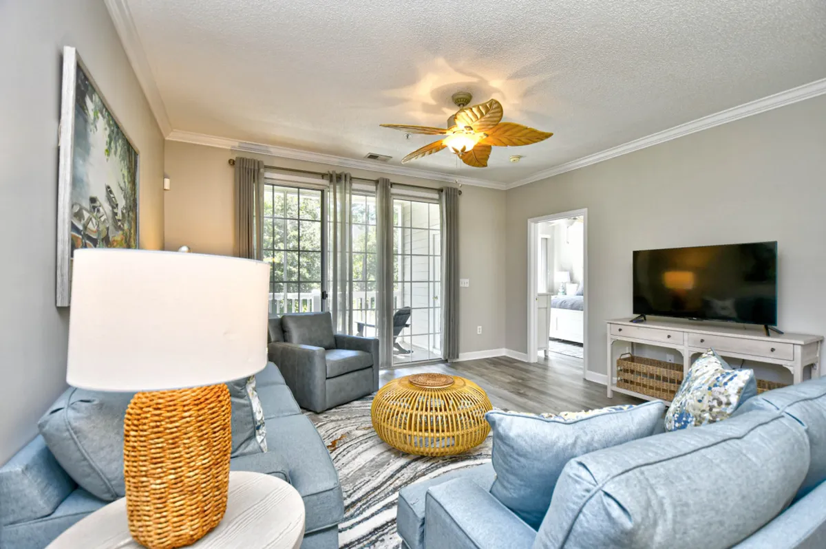 A cozy living room with a plush sofa, ceiling fan, coffee table, and TV, set against soft curtains. The decor is inviting, with touches that encourage relaxation, socializing, and quiet time, making it a true heart of the home.