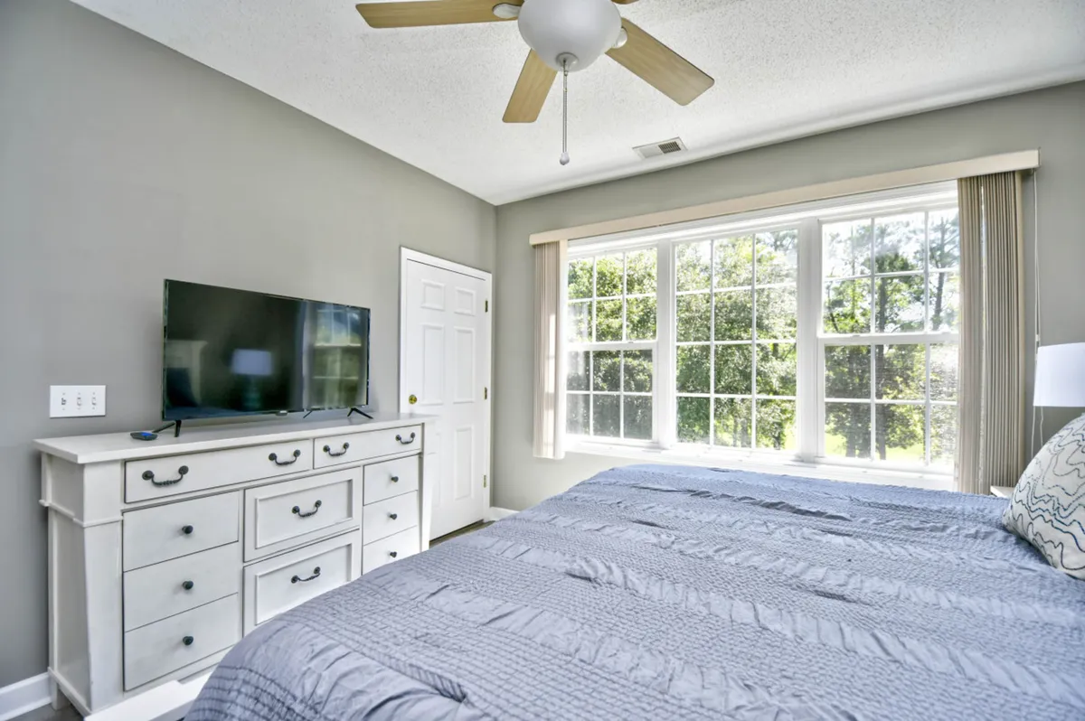 A cozy bedroom scene featuring a bed with textured bedding, a large window allowing in abundant natural light, and a dresser topped with a TV. The space feels calm and inviting, perfect for waking up refreshed or unwinding with a favorite show.