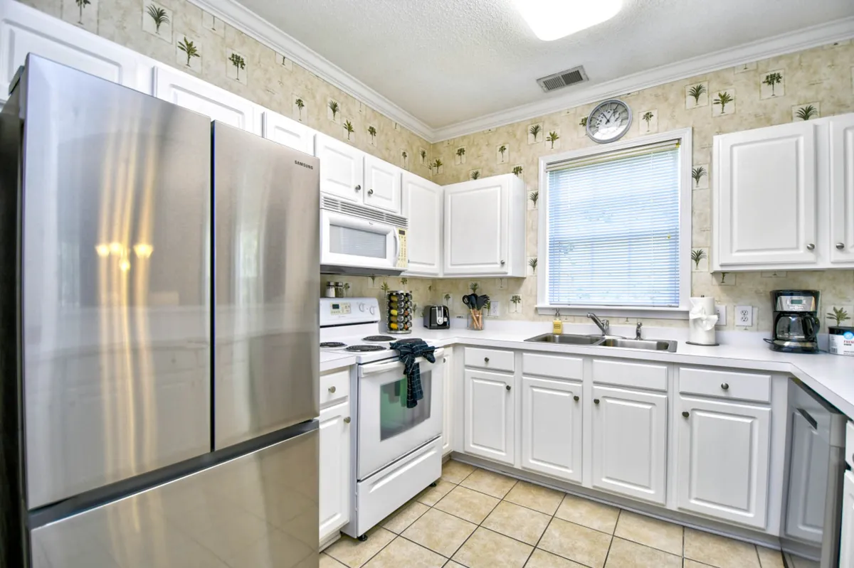 A kitchen view emphasizing a stainless steel refrigerator, gleaming white cabinetry, and natural light streaming in through the window. The open counters and organized design suggest a space ready for culinary creativity and social gatherings.