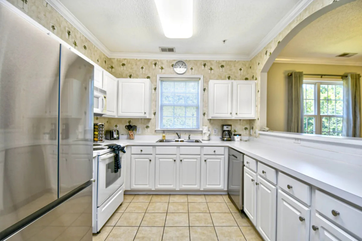 A bright kitchen scene featuring a stainless steel refrigerator, white cabinetry, a window allowing in natural light, a decorative wall, and modern kitchen appliances. The workspace is expansive, with ample counter space and a welcoming layout for both meal prep and gathering.
