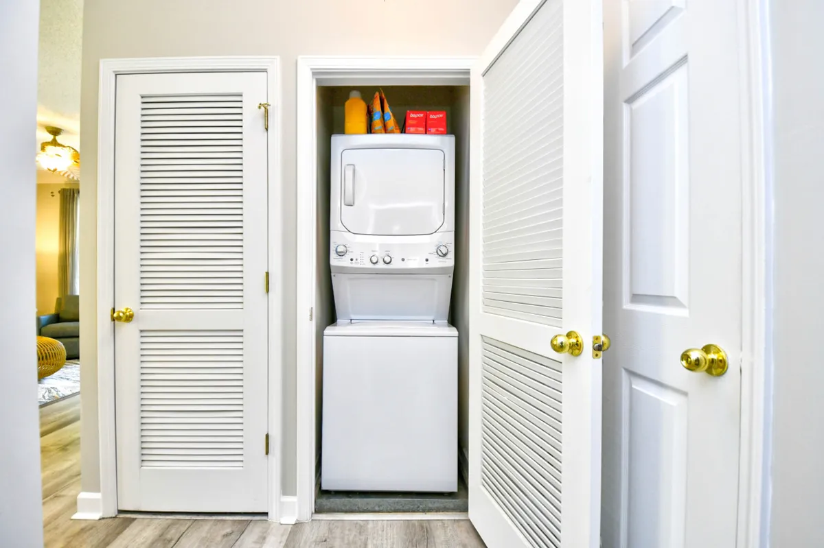 A compact laundry area featuring a stacked washer and dryer, open shelving with laundry supplies, and closet doors. The space is functional and neatly organized, offering convenience for extended stays.