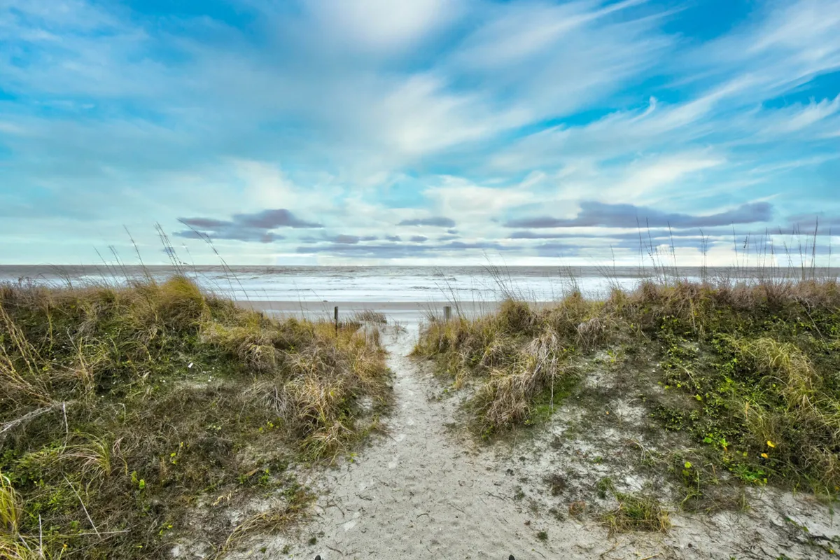A sandy pathway winds through natural dunes and leads directly to the ocean, with gentle waves visible under a serene cloudy sky. The scene evokes both the ease of beach access and the sense of adventure waiting just outside the door.